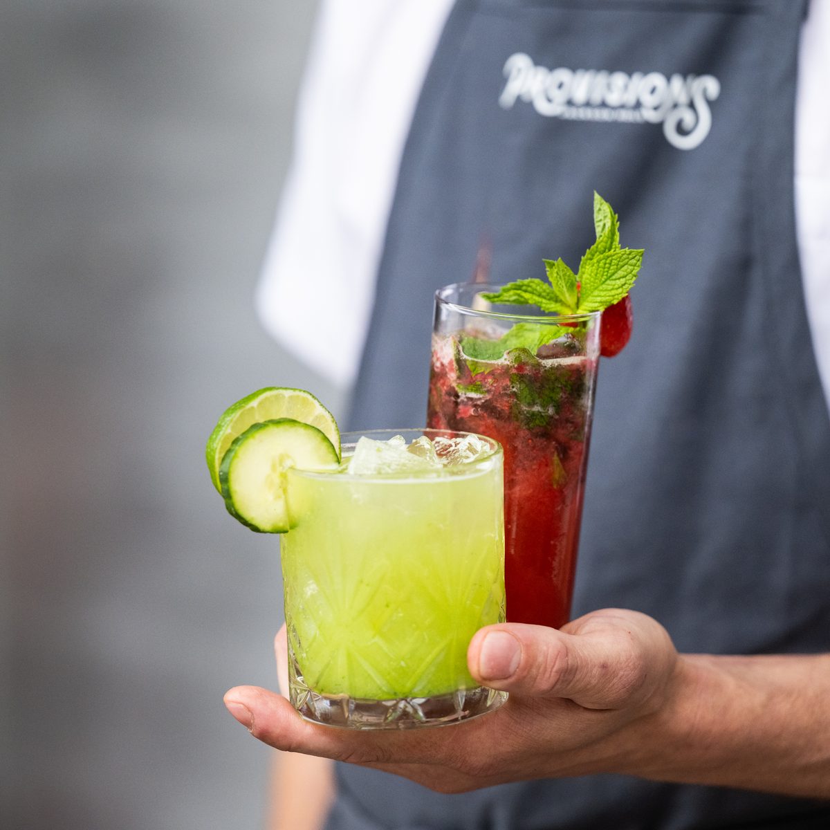 Provisions bartender holding a cucumber cocktail and a berry mint cocktail