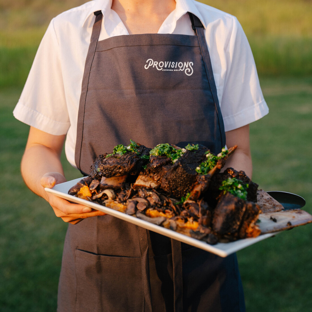 Server carrying a tray of plated dishes through tall grass at a Jackson Hole wedding